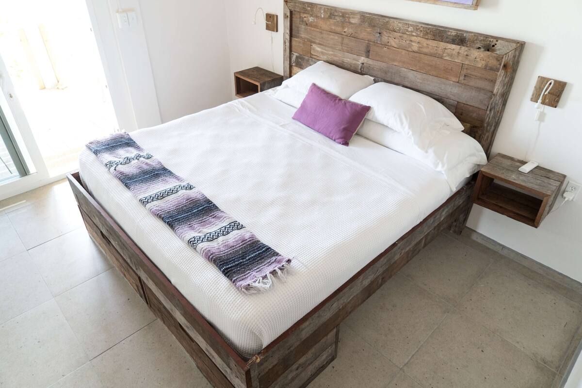 A bed with a wooden headboard, white bedding, a purple pillow, and a patterned throw. There are two bedside tables with lamps and a wall-mounted phone.