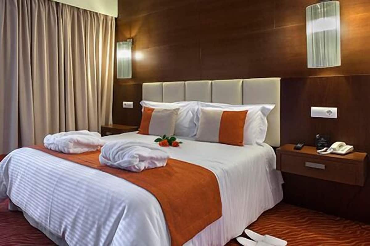 Una habitación de hotel con una cama grande, un cabecero de madera, una mesita de noche con un teléfono y una lámpara, y una ventana con cortinas translúcidas.