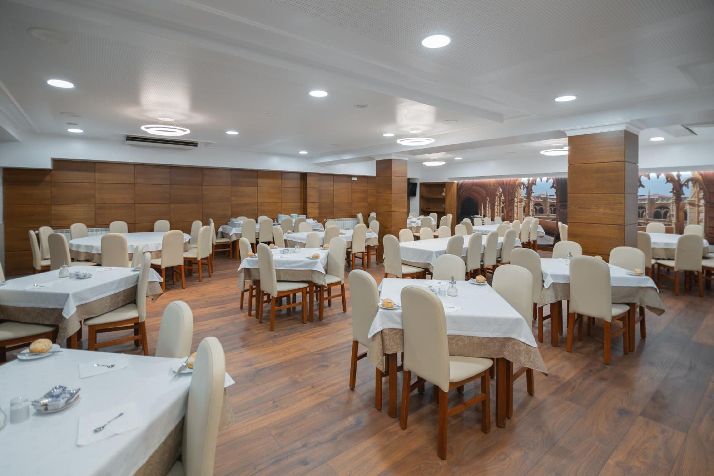 A spacious dining area with multiple tables and chairs, wooden flooring, and a high ceiling with recessed lighting.