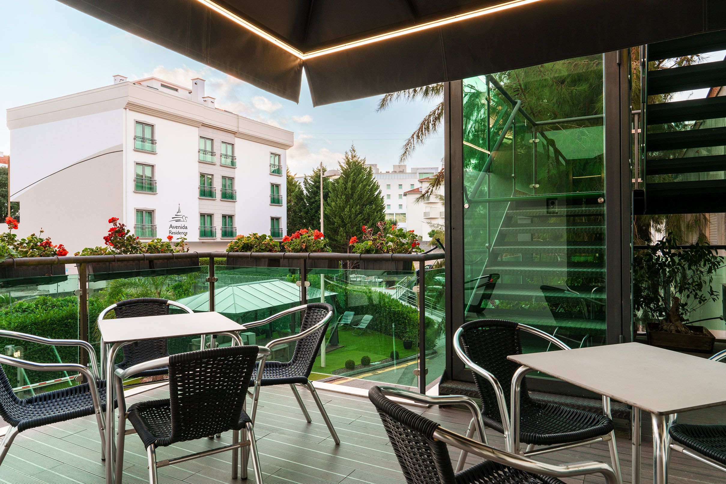 Varanda com área de estar externa, vista de um edifício e guarda-corpo de vidro.