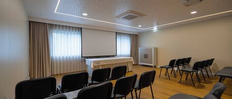 Un salón de conferencias con una pantalla para proyector, una pizarra y varias filas de sillas negras.