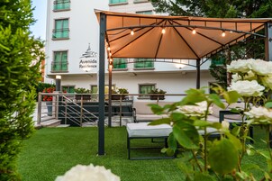 Una terraza en la azotea con una pérgola, mobiliario exterior y vistas a un edificio con balcones.