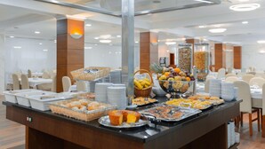 A buffet setup with various food items, including fruits, pastries, and bread, arranged on a counter in a well-lit room with tables and chairs in the background.