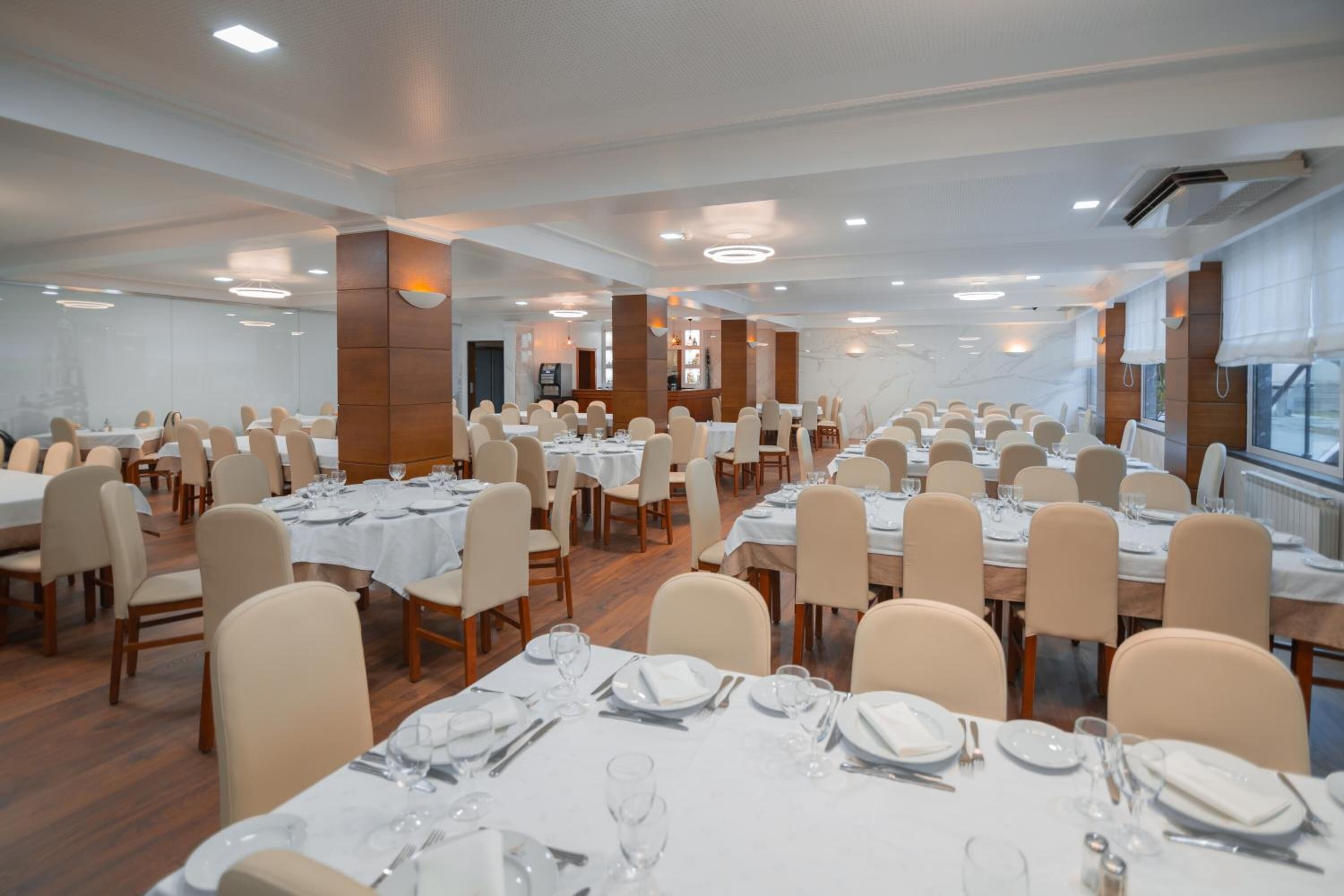 A large banquet hall with multiple round tables, each set with white tablecloths, plates, and cutlery. The room has a modern design with wooden paneling and large windows.