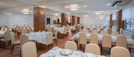 A large banquet hall with multiple round tables, each set with white tablecloths, plates, and cutlery. The room has a modern design with wooden paneling and large windows.