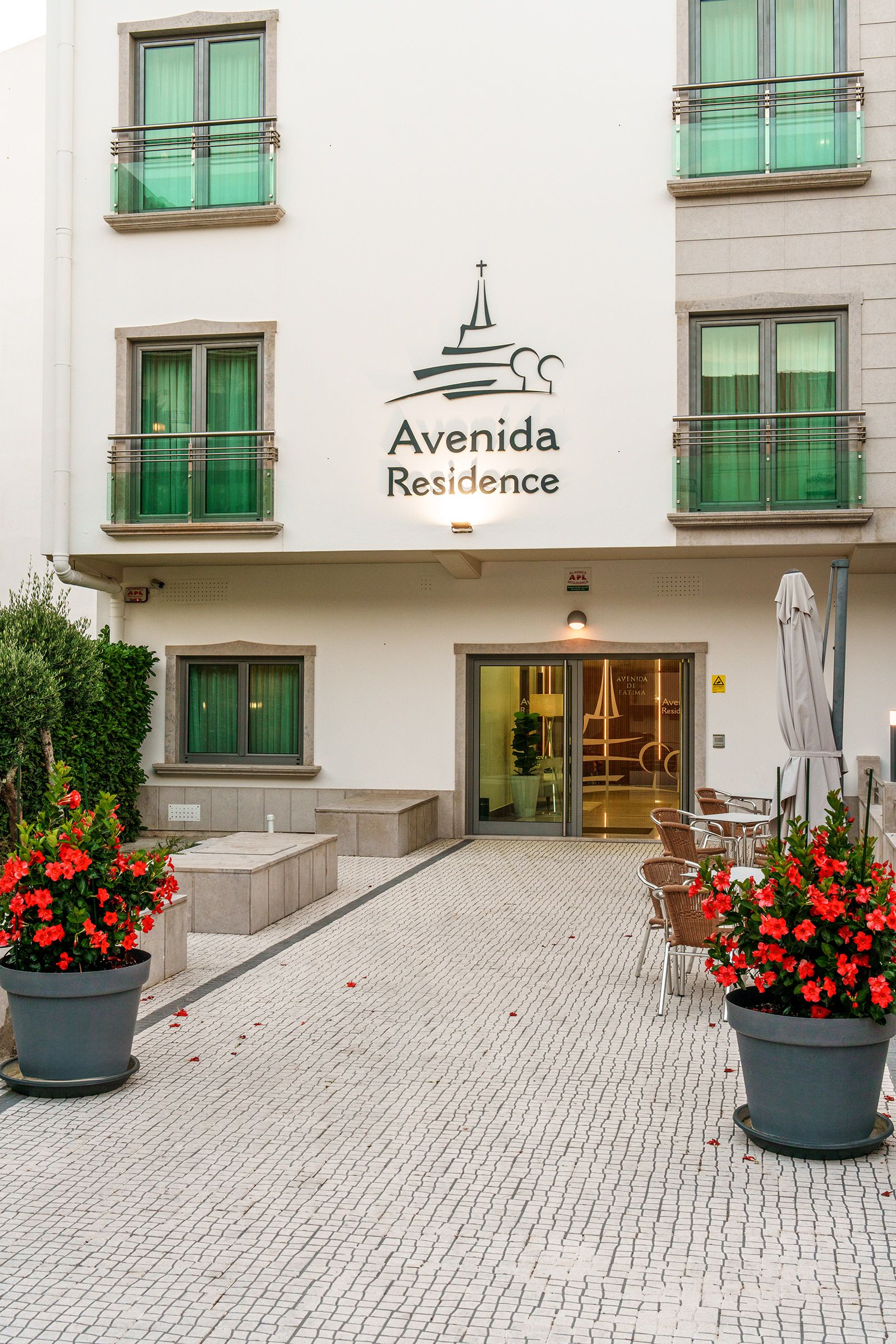 A hotel entrance with a sign reading 'Avenida Residence' and a logo above it. There are potted plants and outdoor seating visible.