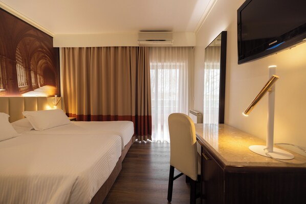 Habitación de hotel con cama, escritorio, silla, televisor y un amplio ventanal con cortinas.