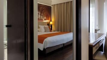Habitación de hotel con cama, escritorio, espejo, lámpara y un cuadro en la pared.