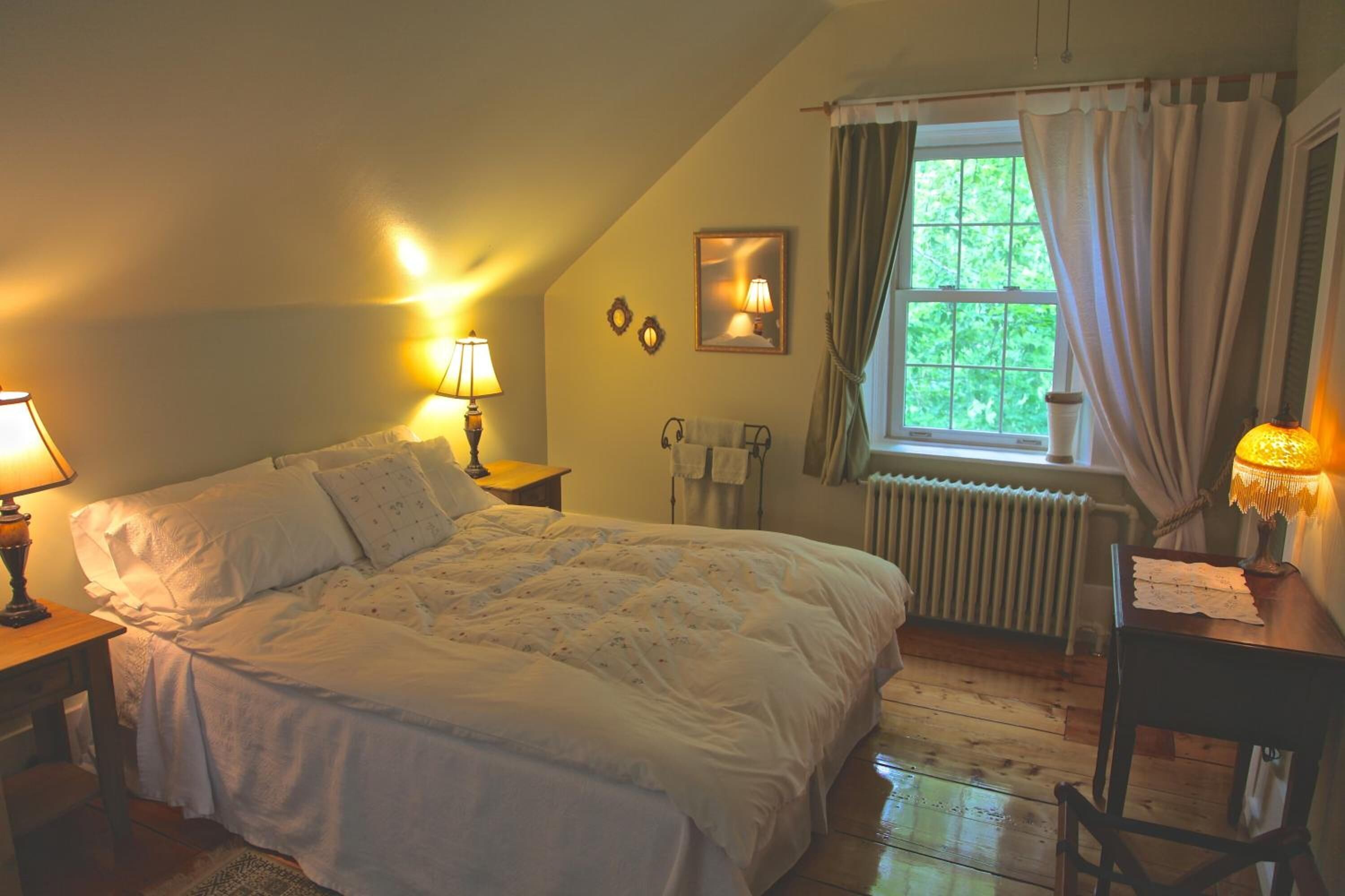 Ett mysigt sovrum med en stor säng, två sänglampor, ett fönster med gardiner, ett element och ett litet bord med en lampa.