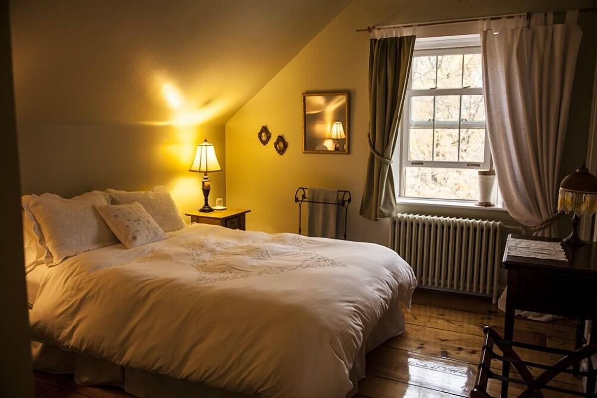 A cozy bedroom with a large bed, a bedside table with a lamp, a radiator, and a window with curtains.