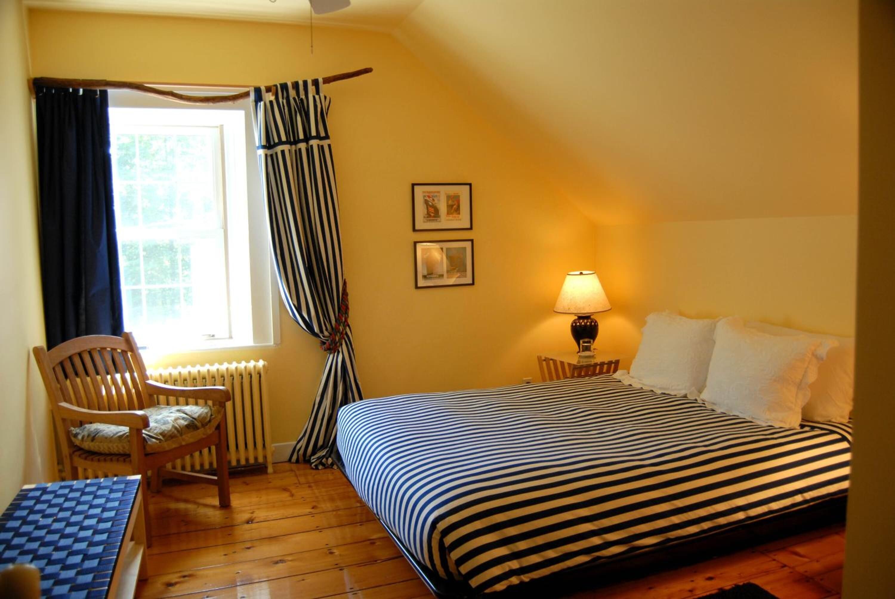 Ein Zimmer mit einem Bett, einem Sessel, einem Tisch, einer Lampe und einem fensterbepflanzten Fenster.
