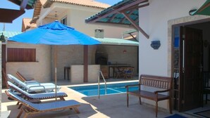 A poolside area with lounge chairs and a bench, a covered seating area, and a brick building with a chimney. - Pousada Casa do Telhado Verde (Cabo Frio)
