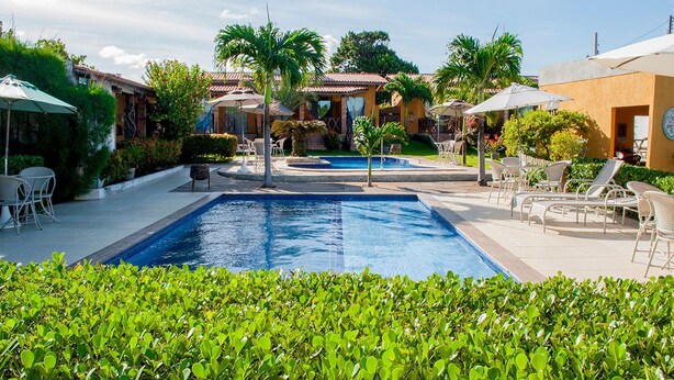 Una piscina con sillones de descanso y sombrillas, rodeada de vegetación y edificios.