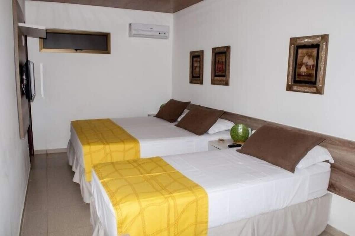 A hotel room with two beds, a bedside table, a wall-mounted air conditioner, and framed pictures on the wall.