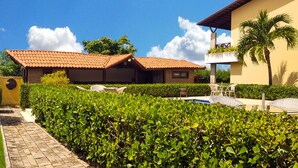 Un jardín bien cuidado con un parque infantil de madera, un porche cubierto de paja y una zona de piscina con sillones de descanso.