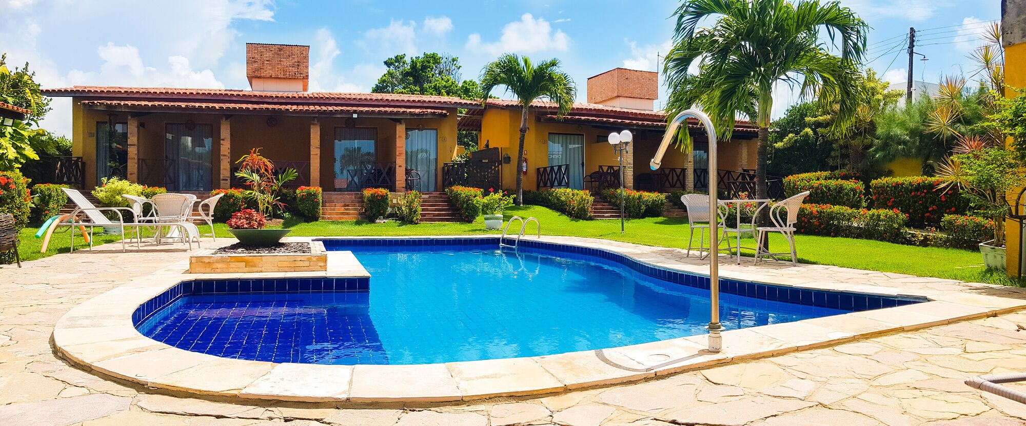 Piscina com deck ladrilhado, rodeada por um jardim bem cuidado e uma casa com telhado telhado.