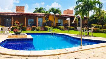 Piscina com deck ladrilhado, rodeada por um jardim bem cuidado e uma casa com telhado telhado.