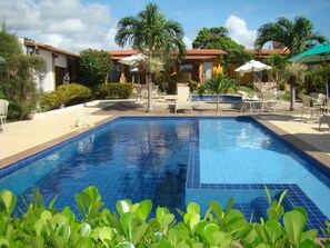 A swimming pool with tiled edges, surrounded by lounge chairs and umbrellas, with palm trees and a building in the background. - Pousada Aconchego (Pitimbu)