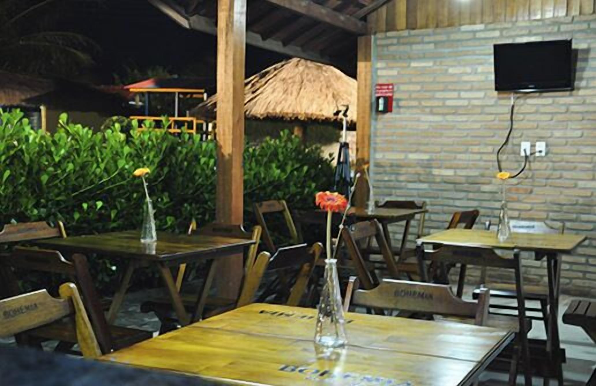 An outdoor dining area with wooden tables and chairs, a thatched roof, and a brick wall with a mounted TV.