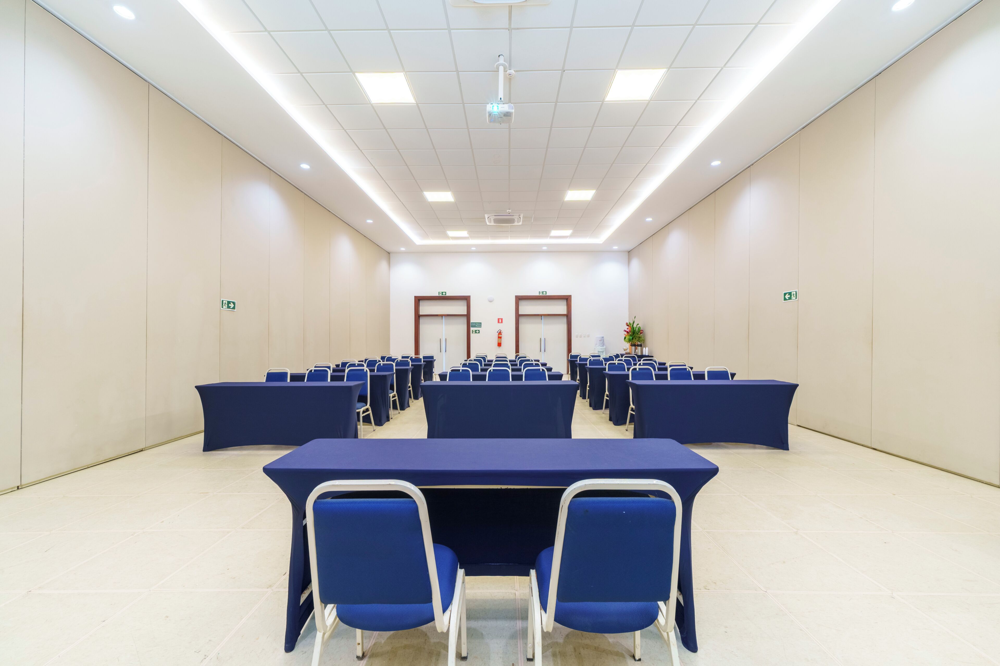 A conference room with rows of tables and chairs, a projector, and emergency exit signs.