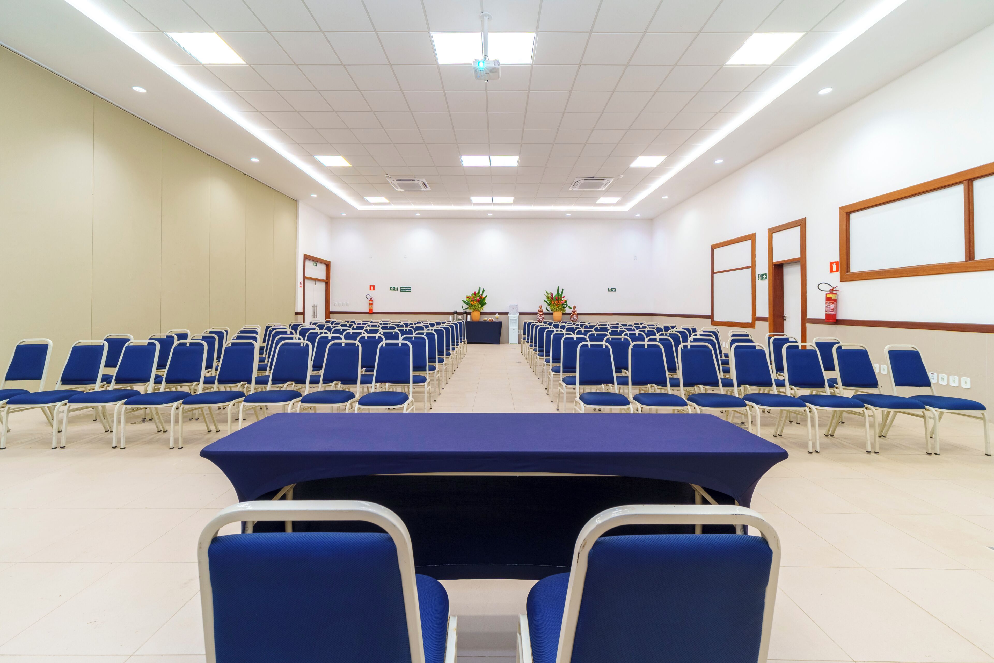 Un salón de conferencias con filas de sillas azules, una mesa larga y una pantalla de proyector al frente.