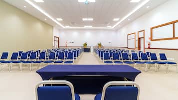 Un salón de conferencias con filas de sillas azules, una mesa larga y una pantalla de proyector al frente.