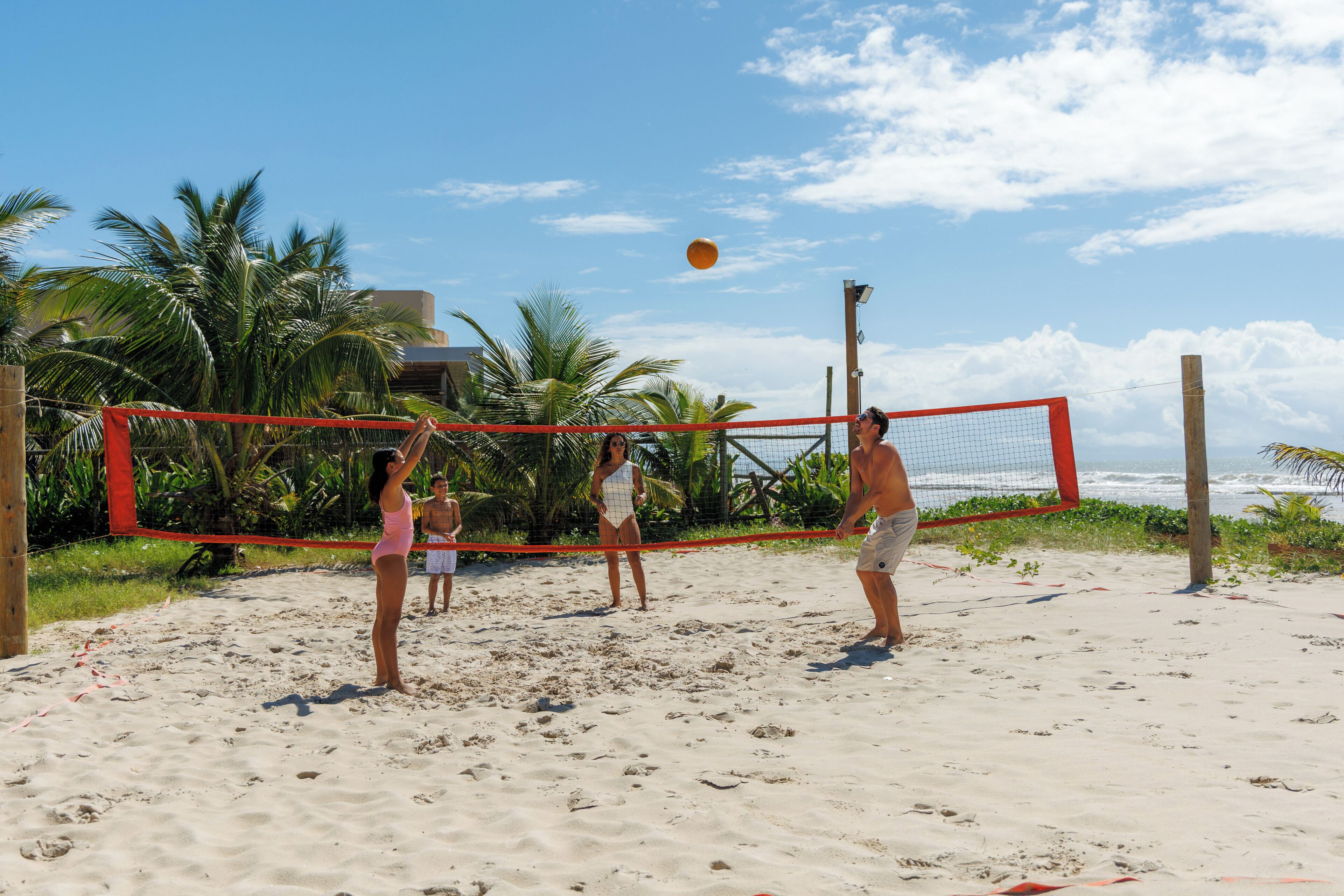 A beach volleyball game with players and a net, palm trees, and ocean in the background.