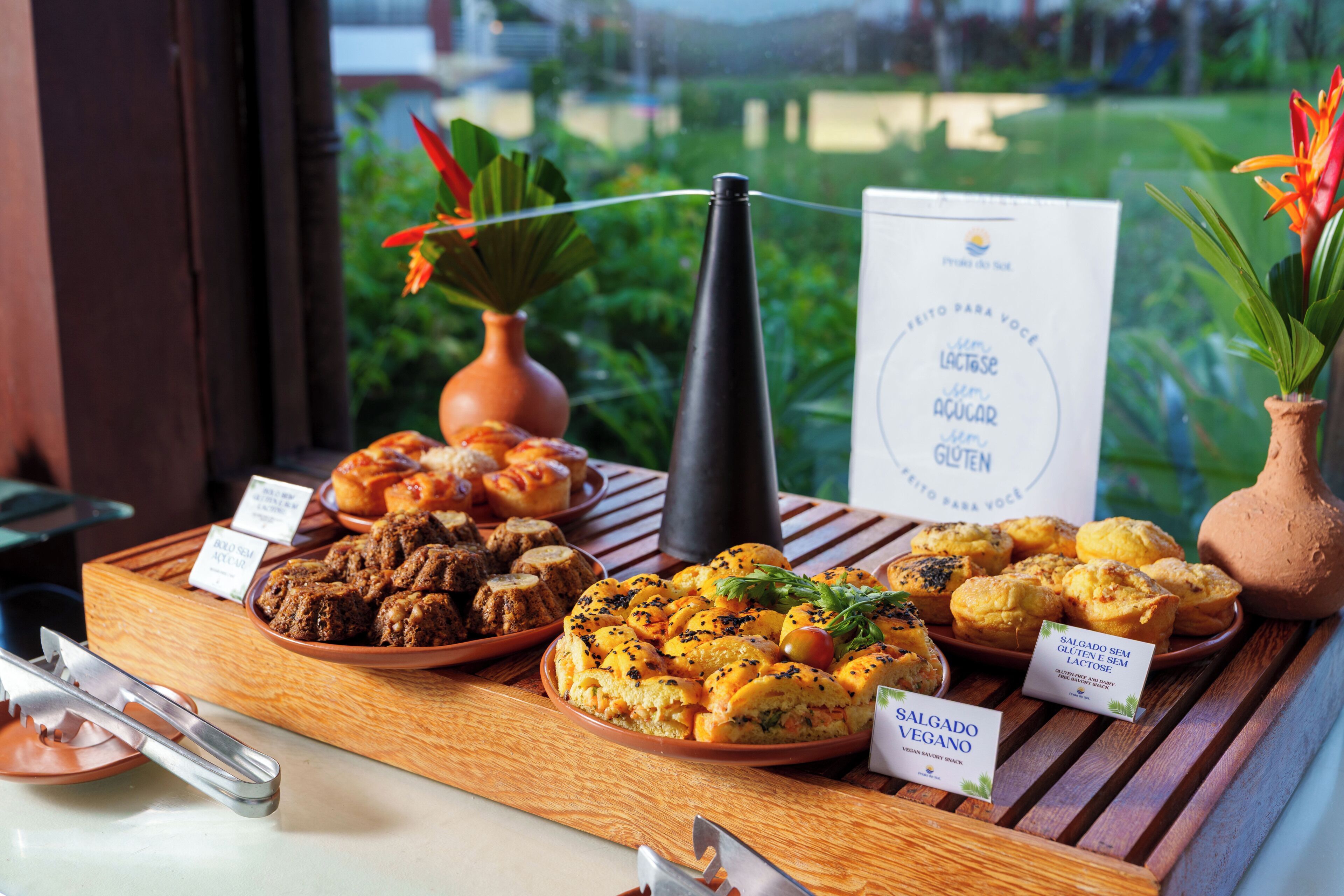 Diversos produtos de panificação sobre uma mesa de madeira, com vista para o verde lá fora.