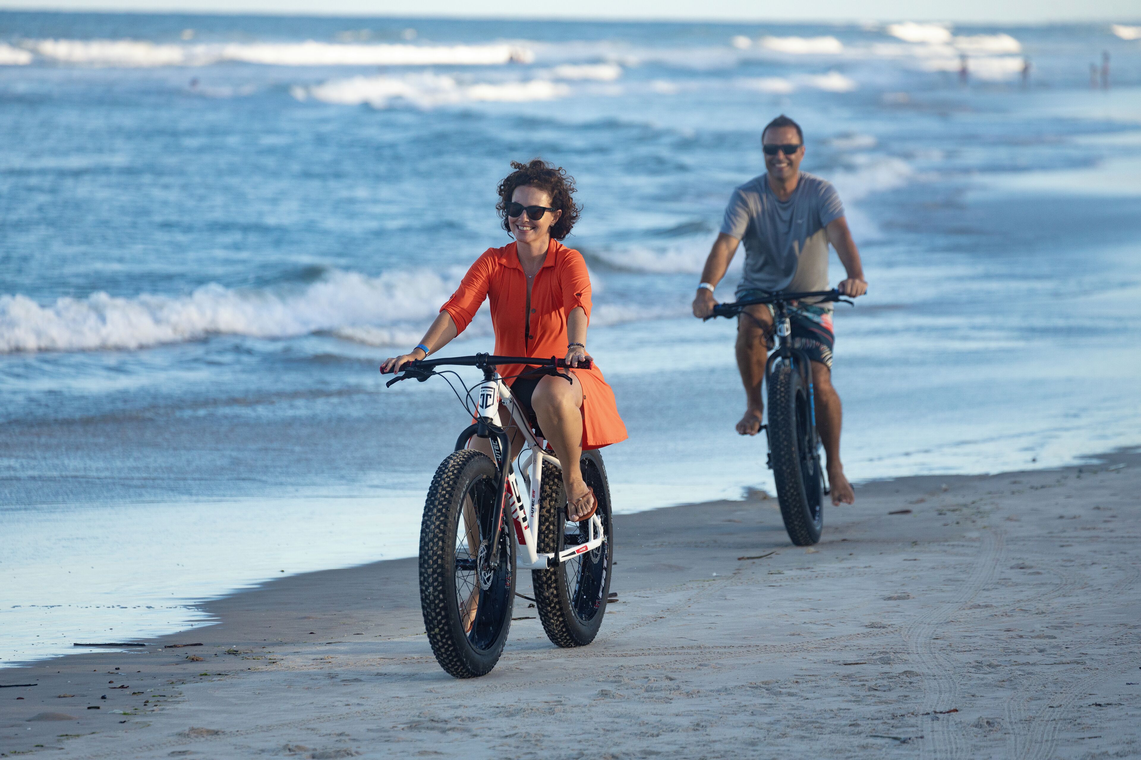 Dos personas andan en bicicleta sobre una playa arenosa con el océano al fondo.