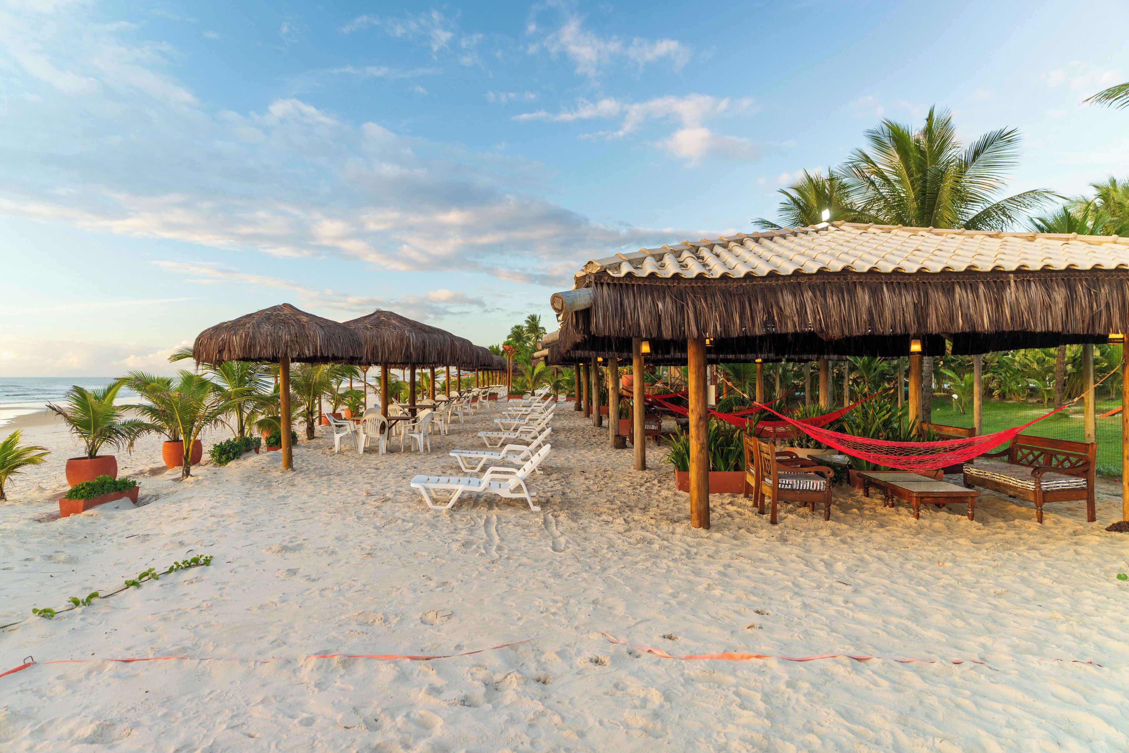 Um resort na praia com estruturas de telhado de palha, cadeiras de lounge e redes.