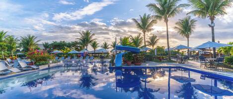 A clear swimming pool with lounge chairs and umbrellas, surrounded by palm trees and a clear sky.