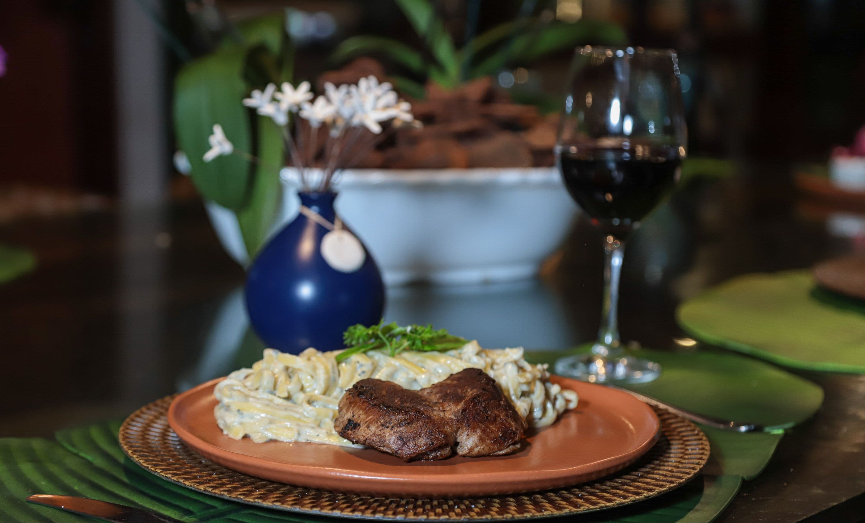 Prato de comida com bife e massa, um copo de vinho tinto, e um vaso azul com flores brancas sobre uma mesa.