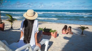 A beach scene with a person sitting on a white chair facing the ocean, two children playing in the sand, and a palm tree in the background.