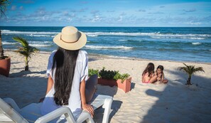 A beach scene with a person sitting on a white chair facing the ocean, two children playing in the sand, and a palm tree in the background.
