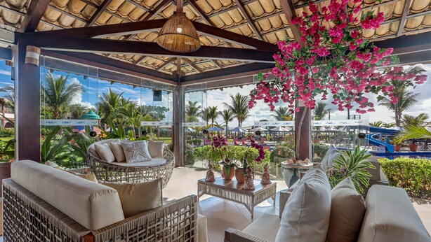 A covered outdoor seating area with wicker furniture, a hanging light fixture, and a view of the pool and palm trees.
