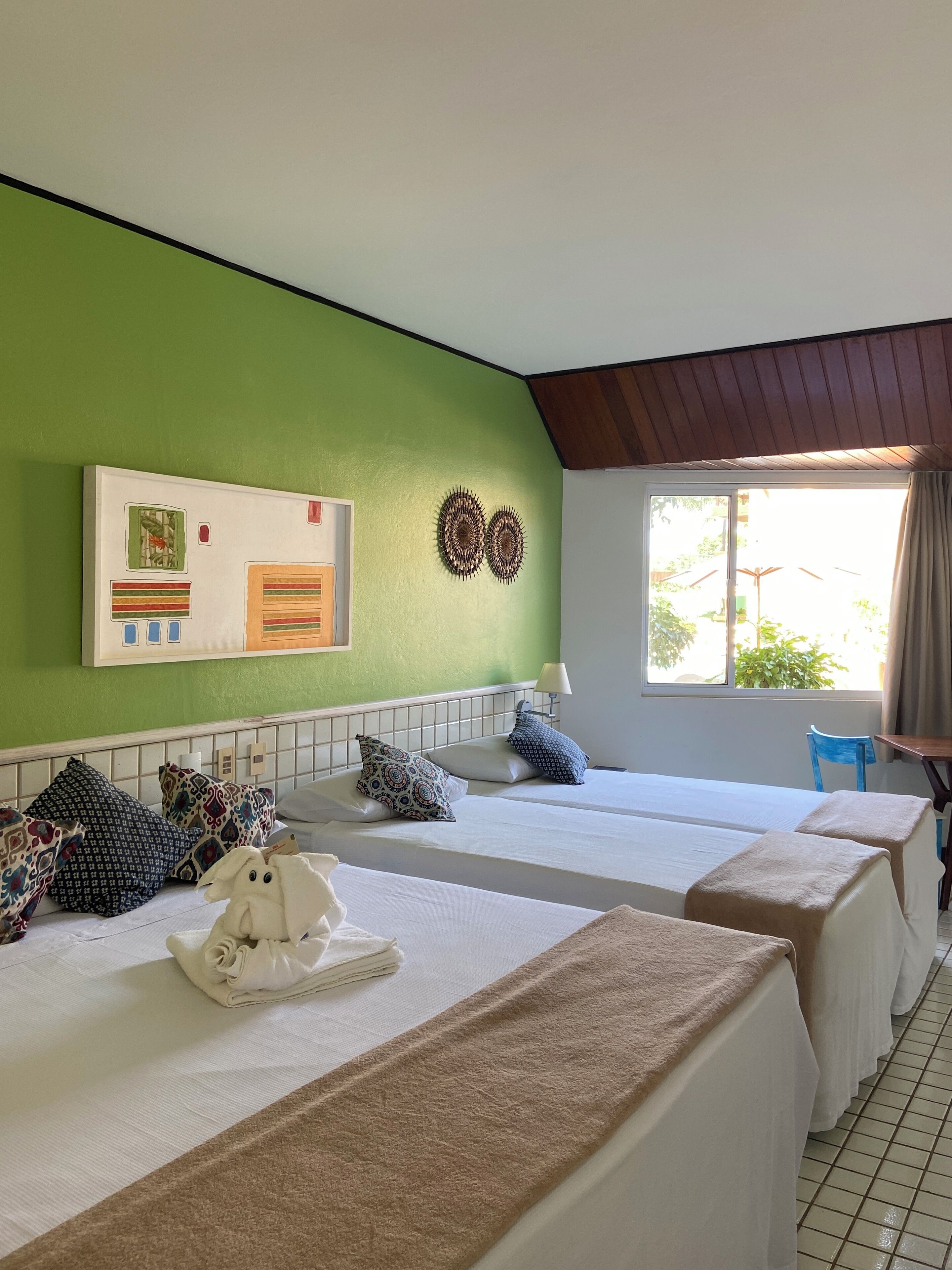 Une chambre d’hôtel avec deux lits, un mur vert, une fenêtre avec des rideaux et une vue sur la verdure à l’extérieur.