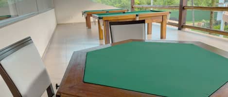 A pool table with a green felt surface, surrounded by chairs and tables in a room with large windows and a view of greenery outside.
