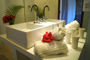Un baño con un lavamanos blanco, dos grifos, una toalla con una flor roja y varias tazas blancas.