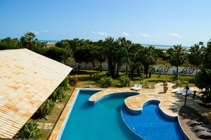 A swimming pool with a curved design, surrounded by lounge chairs and palm trees, with a view of the ocean in the background. - Refúgios Parajuru (Beberibe)