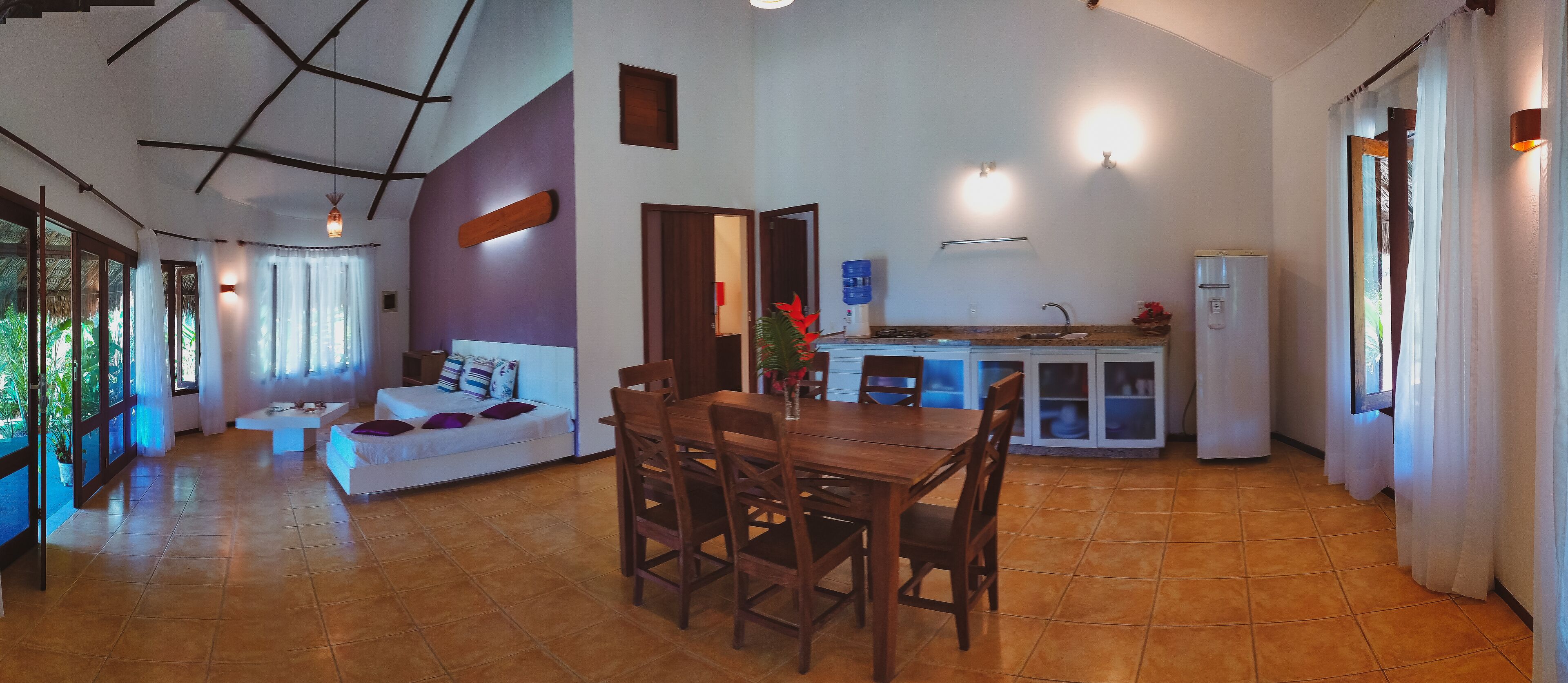 Uma sala de estar ampla, com uma mesa de jantar e cadeiras, um sofá e uma cozinha equipada com geladeira e armários.