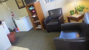 A living room with a black leather chair, a wooden bookshelf, a small table with a lamp, and a window with a view of the outside.