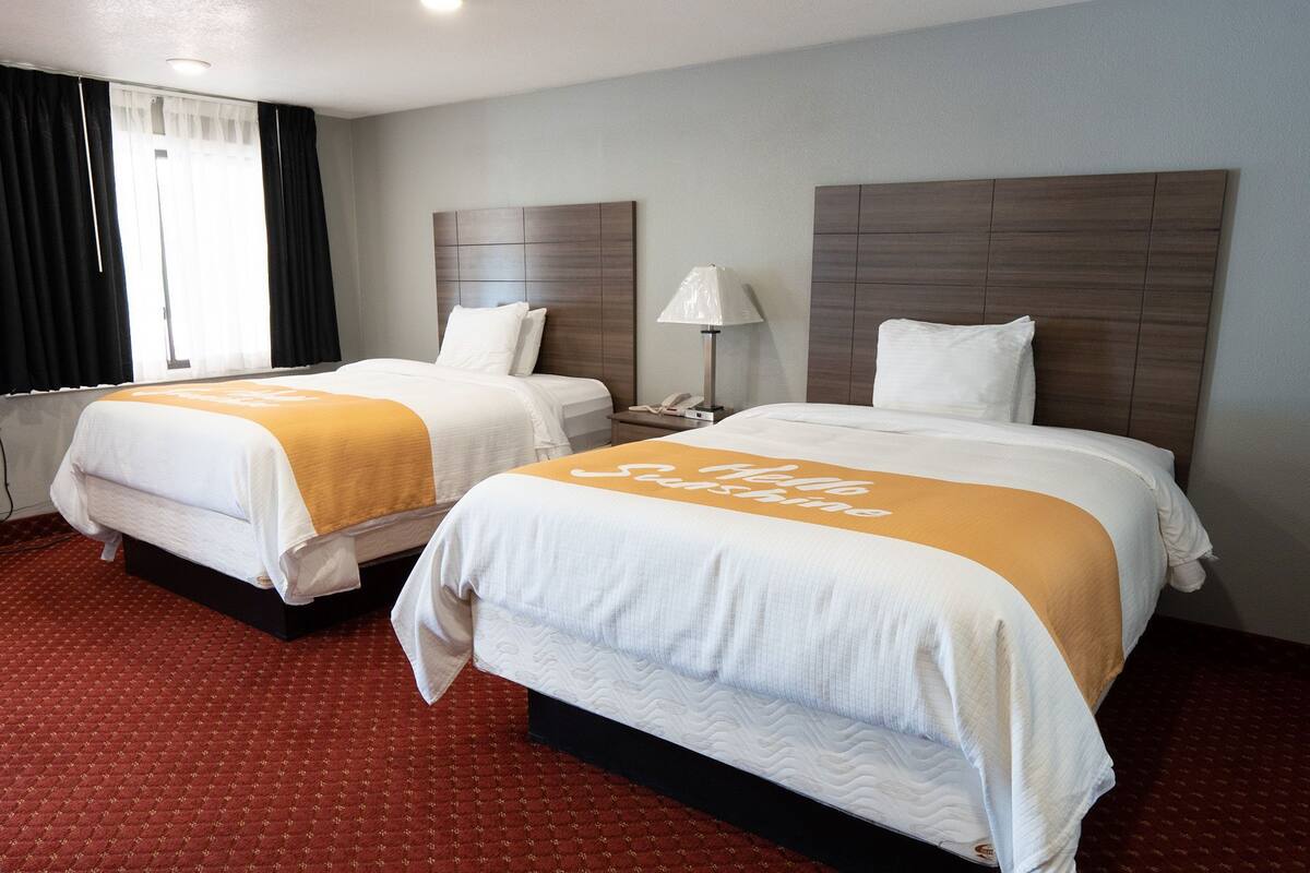 A hotel room with two beds, each with a white and orange bedspread, a nightstand with a lamp, and a window with curtains.