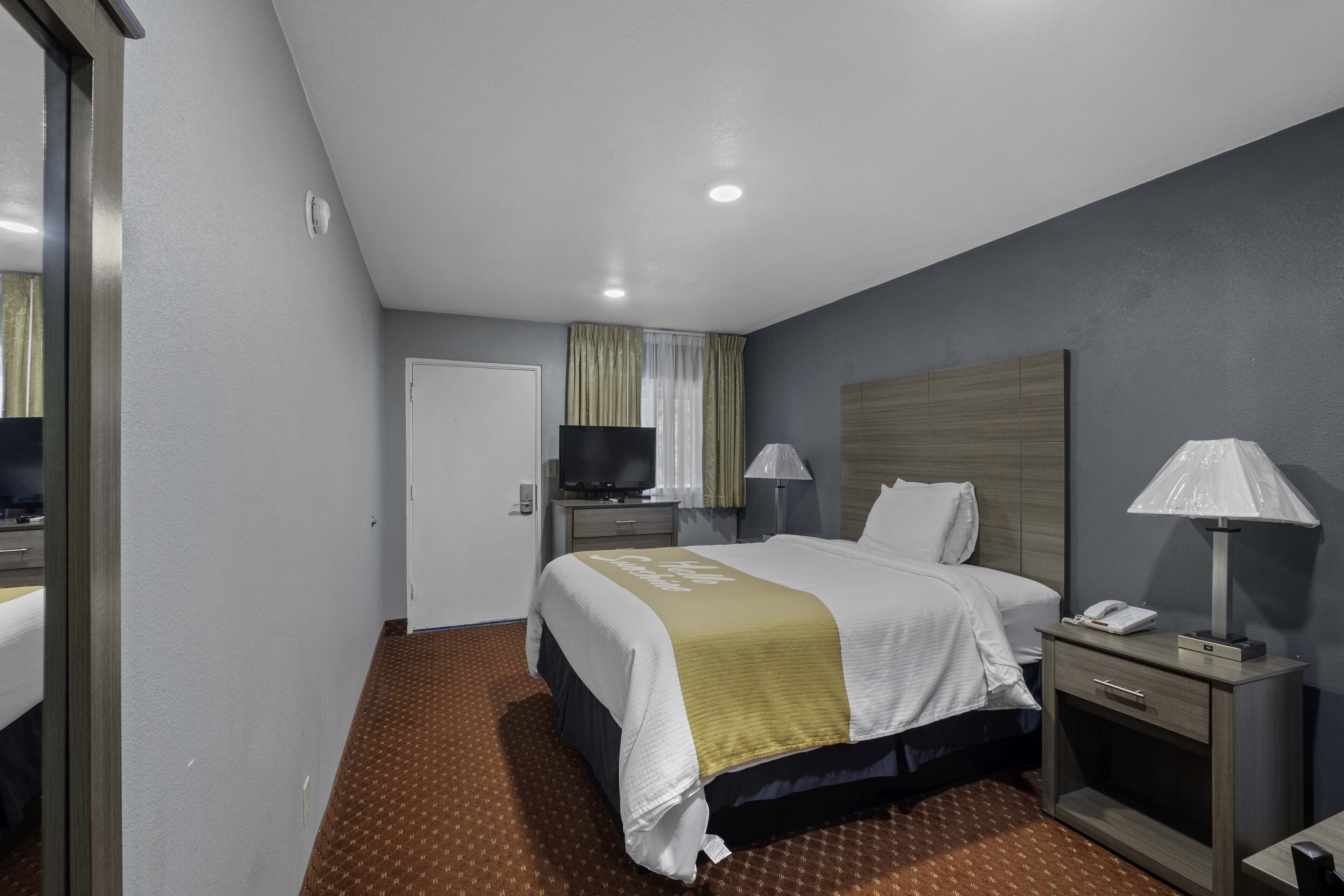 Quarto de hotel com cama, mesa de cabeceira, abajur e televisão.