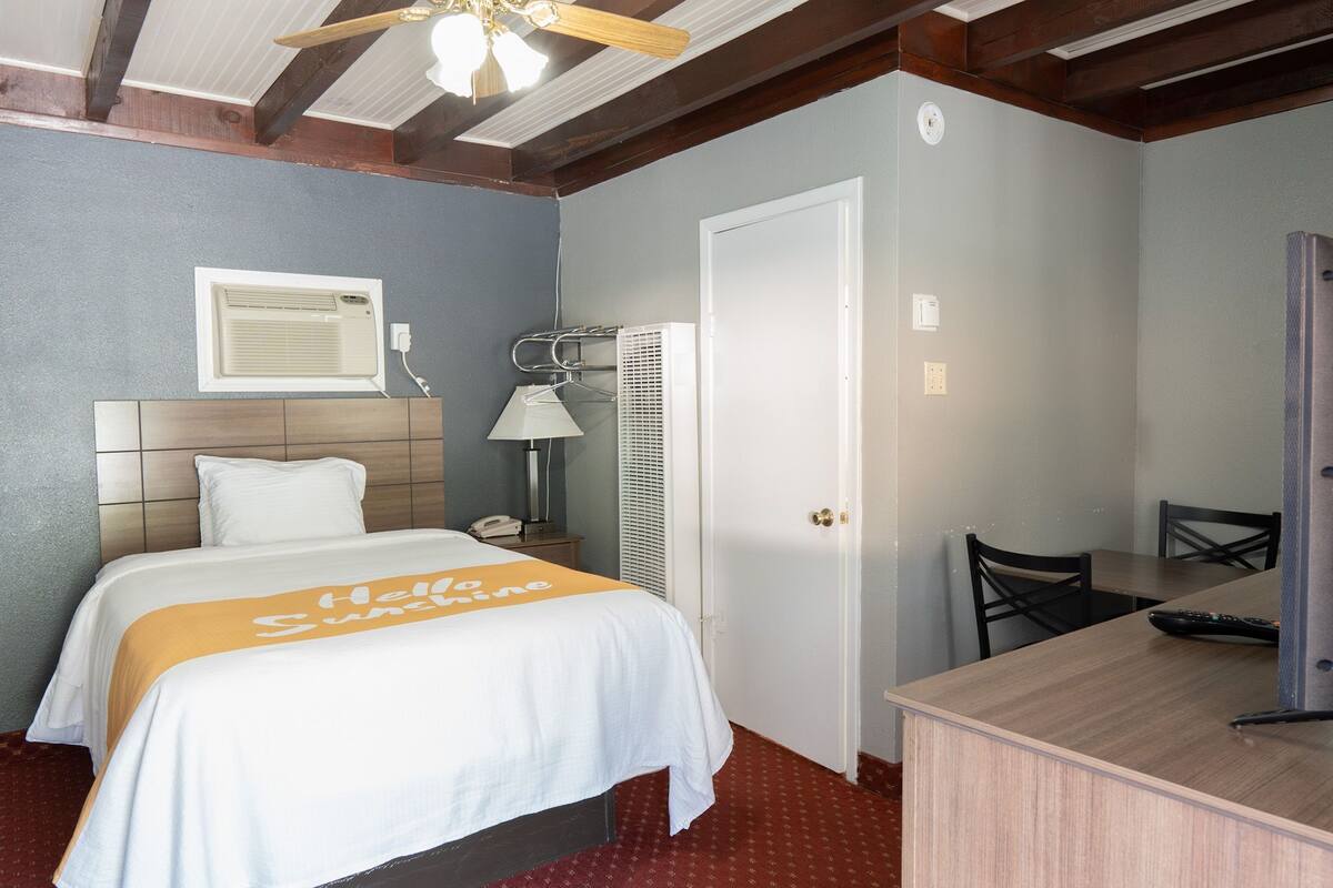 A hotel room with a bed, a desk, a chair, a ceiling fan, and a door.