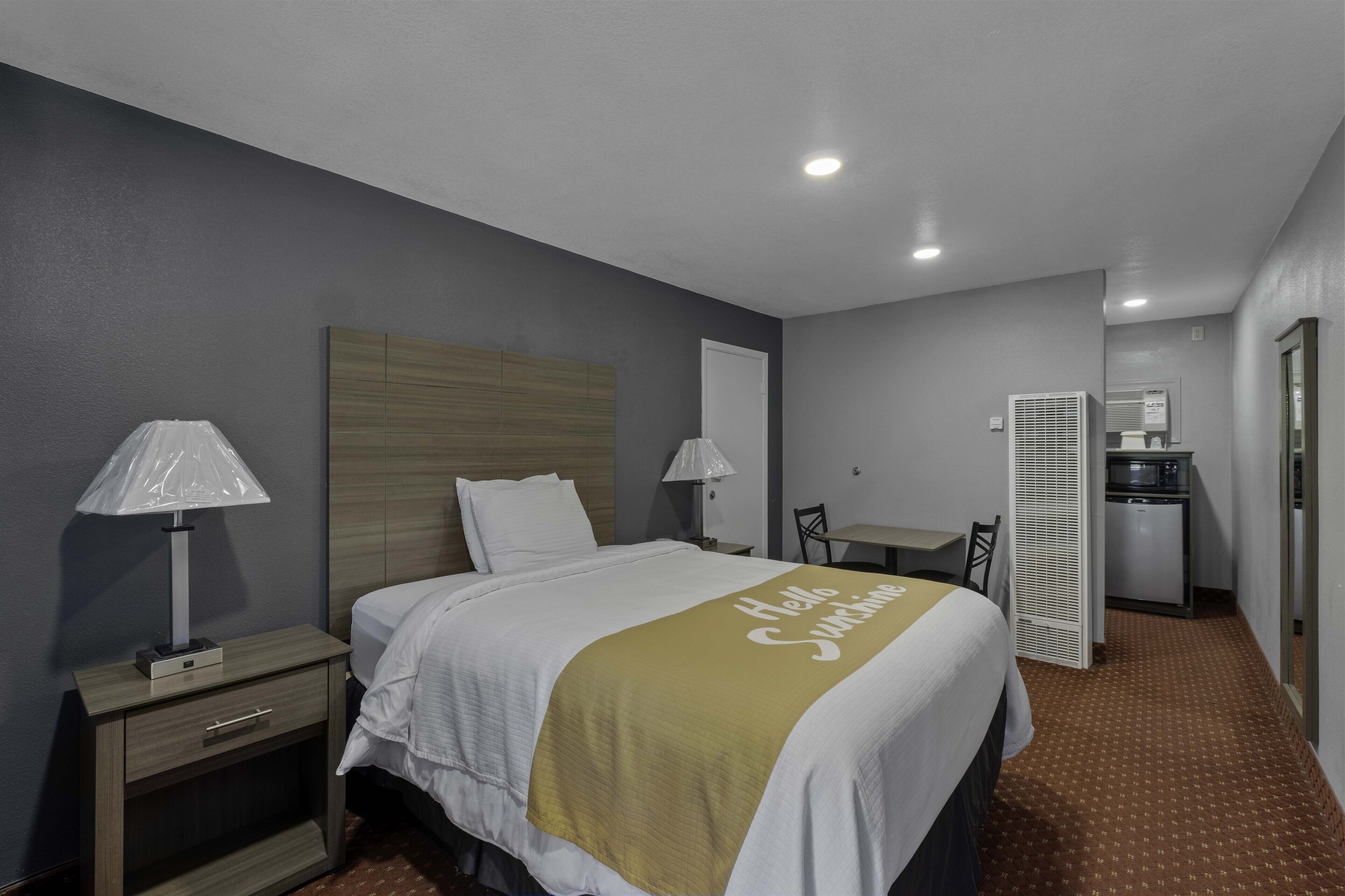 Una habitación de hotel con cama, lámparas de noche, una mesita, una silla y una kitchenette.