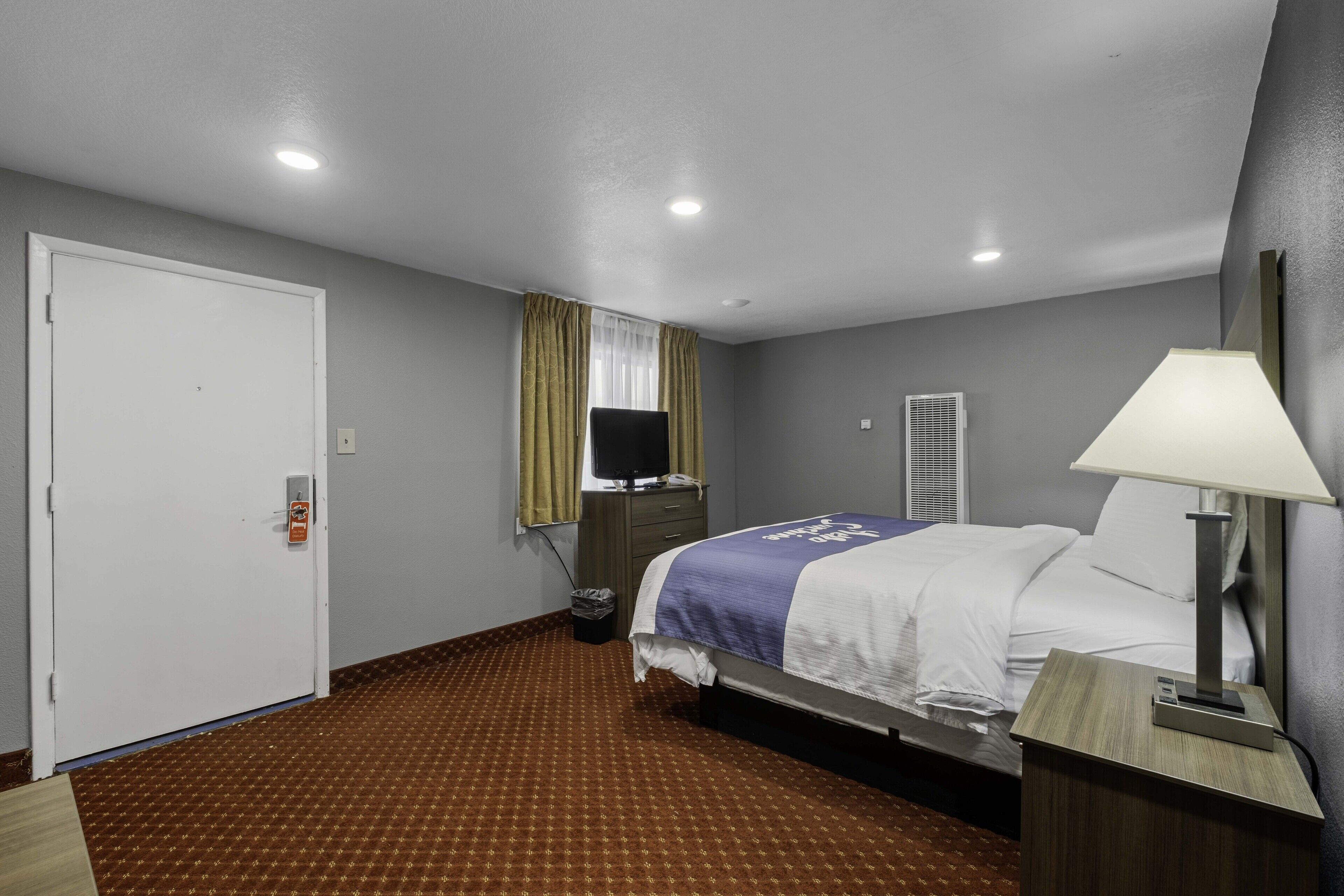 A hotel room with a bed, a desk, a lamp, a TV, and a window with curtains.