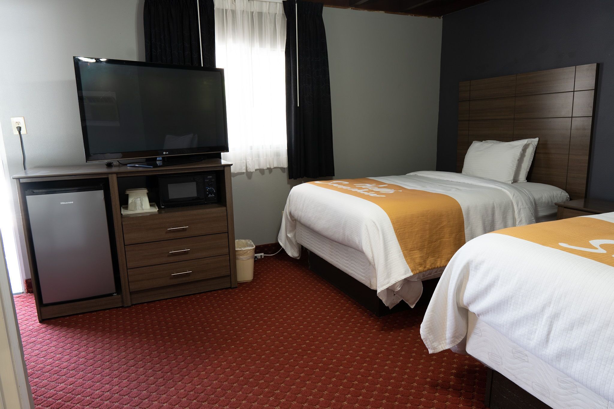 Une chambre d’hôtel avec un téléviseur posé sur un meuble, un bureau avec un ordinateur, deux lits avec une literie blanche et orange, et un sol recouvert d’une moquette rouge.