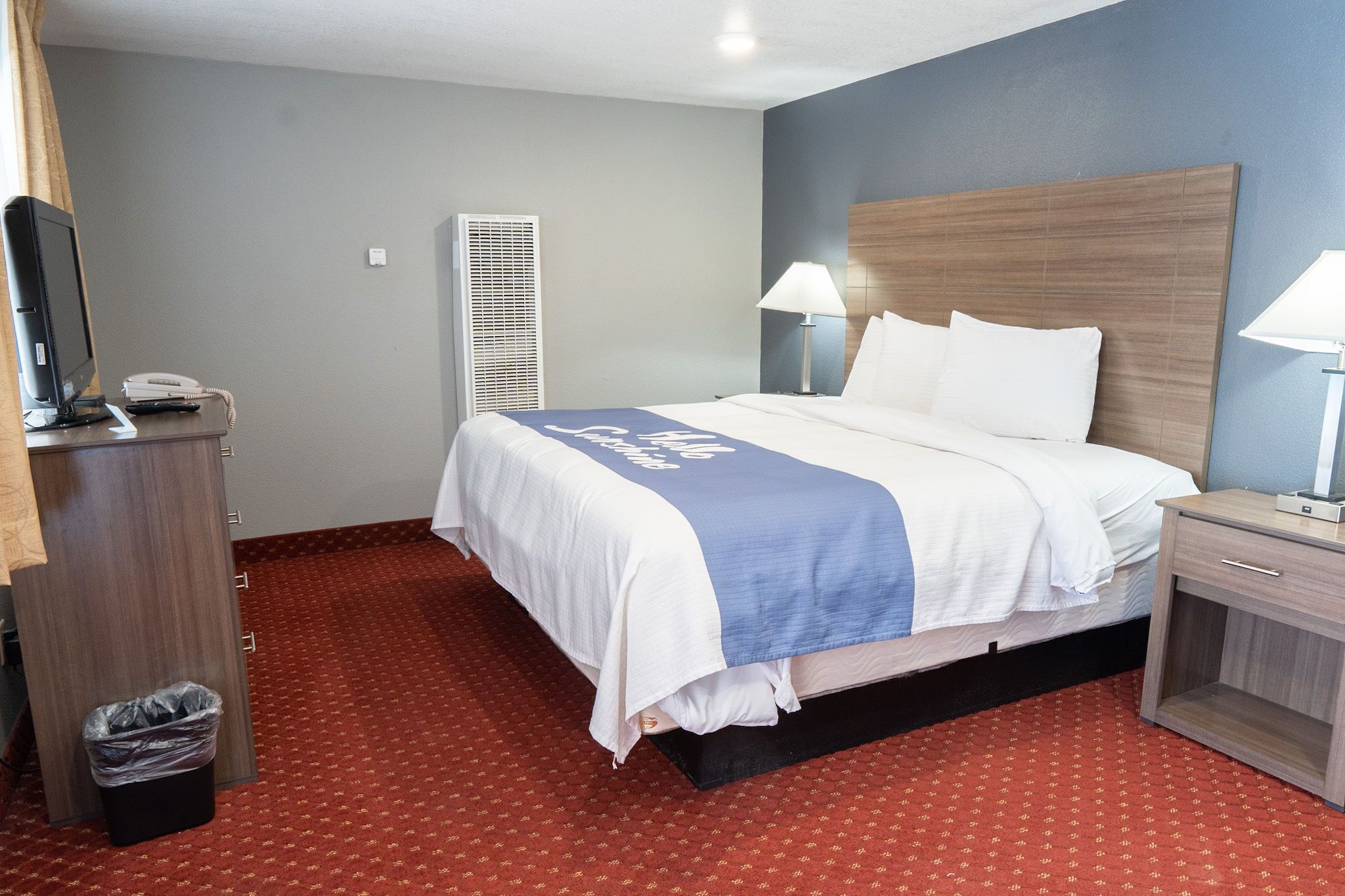 A hotel room with a bed, bedside lamps, a desk with a telephone, and a trash bin.