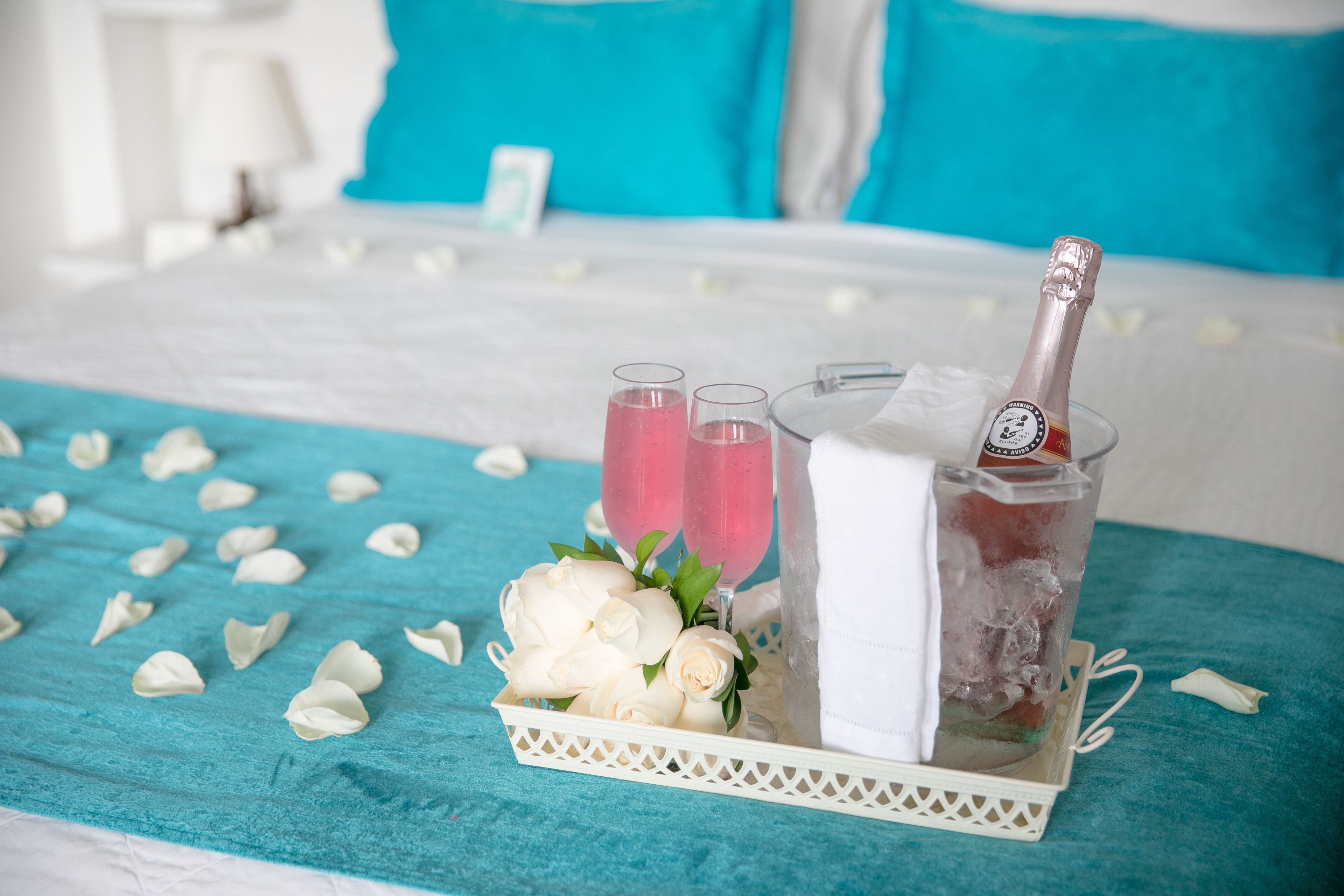Un lit recouvert d’une couvre-lit turquoise, un plateau avec une bouteille de champagne, des verres et des roses blanches.