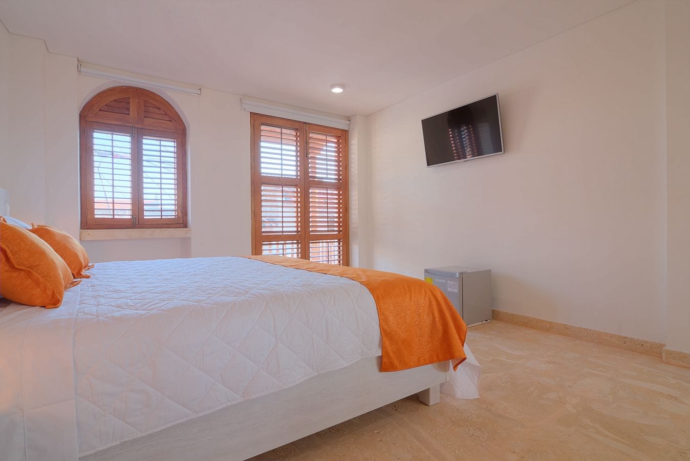 Un dormitorio con una cama grande, dos ventanas con contraventanas de madera, un televisor de pantalla plana montado en la pared y un equipo de aire acondicionado pequeño.